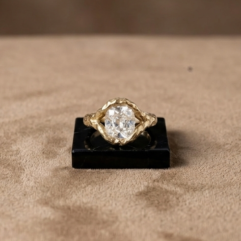 A yellow gold ring with a large clear round-cut gemstone, displayed on a small black stand against a textured beige background.